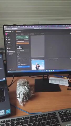 The video shows a desktop computer setup with two monitors, a laptop, and a keyboard on a wooden desk. The main monitor displays a dark interface with various menus and text in Russian, likely a software application. The laptop, positioned to the left, also shows a dark screen with some lines of text.

A clear plastic bag containing small metal objects, possibly screws or hardware, sits on the desk between the laptop and the main monitor. A woman is partially visible in the background, looking towards the right side of the frame, with her arm resting near her face.

The scene appears to be indoors, with a white ceiling and a white lamp hanging overhead. The lighting suggests it could be daytime, but there are no external windows visible to confirm the weather or time of day. The overall impression is that of a workspace, possibly an office or a home study area.

The visible text on the main monitor includes words like "Magnifico" and "Тестирование," suggesting a testing or review process. The laptop screen has "HUAWEI" branding. There's also a small image of a smiling woman in business attire on the main monitor.

The user is interacting with the computer system, as indicated by the dynamic content on the screens. The presence of the hardware suggests a task involving assembly, repair, or a project that requires such components.