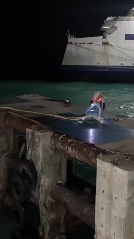 A lone person, wearing a white hard hat and an orange high-visibility vest, stands on a concrete pier at night. They are hunched over, holding a flashlight, which illuminates the ground around them. The person appears to be engaged in some kind of work or inspection.

In the background, a large white ferry is docked. Its lights are on, casting a glow on the dark water. The sea is a deep turquoise, reflecting the faint light. The pier itself is industrial, with thick, rusty pipes and thick mooring ropes visible in the foreground. Several old tires are attached to the structure, likely as fenders.

The scene is dark, suggesting it is late at night. There are no visible signs of other people or activity. The overall impression is one of solitary work in a port environment. The location cue of Nathon, Thailand, places this scene in a tropical setting, though the weather cannot be determined from the image alone. No text is visible in the media file.
