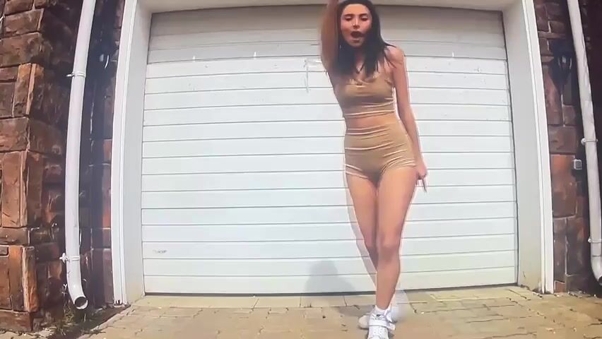 A woman with dark hair is dancing in front of a white garage door. She is wearing a tan crop top and matching shorts, along with white sneakers. The garage door is a roll-up style, and there is a brick wall on the left side of the frame. The ground is paved with interlocking bricks. The woman's movements are energetic, and she appears to be enjoying herself.