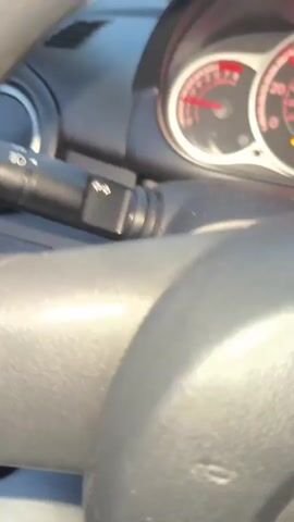 The video shows a view from a vehicle, likely a car, driving down a road with a rainbow visible in the sky. The footage is blurred due to motion. In the foreground, parts of the vehicle's interior are visible, including what appears to be a steering wheel or dashboard. As the vehicle moves, it passes other vehicles on the road and trees lining the side. Traffic lights are visible in the distance, some of which are illuminated. The sky is a pale purple, suggesting it is either dusk or dawn. Another car, dark blue, drives past the camera car.