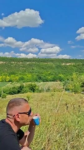 A man with short hair and sunglasses is drinking from a small blue and white cup in a grassy field. He is wearing a black t-shirt. Behind him, there is a lush green hillside under a bright blue sky with fluffy white clouds. The scene is illuminated by natural sunlight.