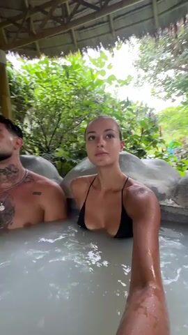 The video shows a man and a woman in a milky white water bath. The woman is wearing a black bikini top and has her hair in a bun. The man is shirtless and has tattoos on his chest and neck. They are both looking to the right. The bath is surrounded by stone-like structures and lush green plants. Above them is a thatched roof structure.