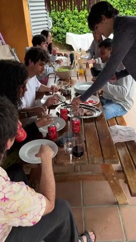 A group of approximately ten young adults are seated around a long wooden picnic table laden with food and drinks. The table is arranged with white plates, cans of Estrella beer, glasses, and platters of grilled meat and sliced tomatoes. A bowl of salad is also visible. People are serving themselves food, passing plates, and talking. A person wearing a gray and black striped long-sleeved shirt is distributing slices of meat onto plates, while another person places tomato slices on a plate. The table is set on a tiled patio with an exterior wall behind the group. The overall atmosphere suggests a casual outdoor gathering, possibly a barbecue or a lunch.