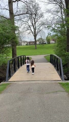 Two young girls are walking across a wooden bridge in a park. The bridge has black metal railings. One girl has long dark hair, a white t-shirt, and blue jeans. The other girl also has long dark hair and is wearing a light-colored shirt and dark pants. They are walking away from the camera toward a green grassy area with scattered trees and houses in the background. The sky is overcast.  Trees line the path leading up to the bridge.