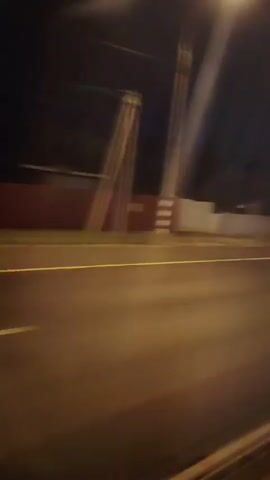The video shows a blurry, nighttime view from a vehicle moving along a road. Streetlights cast long shadows and illuminate the asphalt. A white barrier runs along the side of the road, separating it from a dark, indistinct background that may contain trees or buildings. The motion blur suggests the vehicle is traveling at a moderate speed. There are no visible people or other vehicles in the frame. The overall impression is of a quiet, solitary journey through a suburban or highway environment after dark.