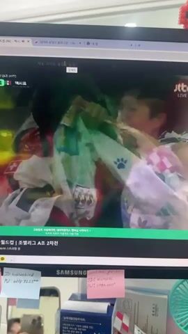 The video shows a computer monitor displaying a soccer match. A male soccer player in a purple jersey with the number 10 on the back and a South Korean flag patch on his left sleeve is visible on the screen. He appears to be reacting to something off-screen with a strained facial expression, possibly anger or frustration. He is looking to his right. Another player, wearing a black jersey with the name "SALAH" and the number 9 on the back, is partially visible in the foreground, facing away from the viewer and towards the first player. The background on the screen shows a blurred green and yellow pattern. Text overlays indicate the match is between South Korea and Mexico, with the score currently at 1-0 to Mexico. Below the screen, the top of a white Samsung branded object is visible, along with colorful sticky notes attached to the monitor's bezel.