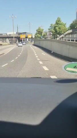 The video shows a view from inside a car driving on a highway. The road has multiple lanes marked with white dashed lines. In the foreground, the dashboard of the car is visible, including the passenger side of the dashboard and the air vent. On the windshield, there is a circular green sticker with an image of a cigarette and a slash through it, indicating no smoking. Ahead, other cars are driving on the road. On the right side of the road, there is a concrete barrier and a metal railing, with trees and foliage visible above. On the left side of the road, there are overhead signs and traffic lights. The sky is clear and blue.