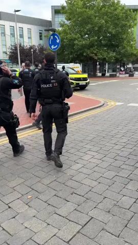 A group of police officers in uniform are walking on a paved area with yellow lines. One officer in the foreground is walking away from the camera, wearing a black uniform with a vest that says "POLICE 13". In the background, several other police officers are standing near two police cars, one of which has its door open. A roundabout sign is visible in the background. The environment appears to be an urban or town setting with buildings and trees.