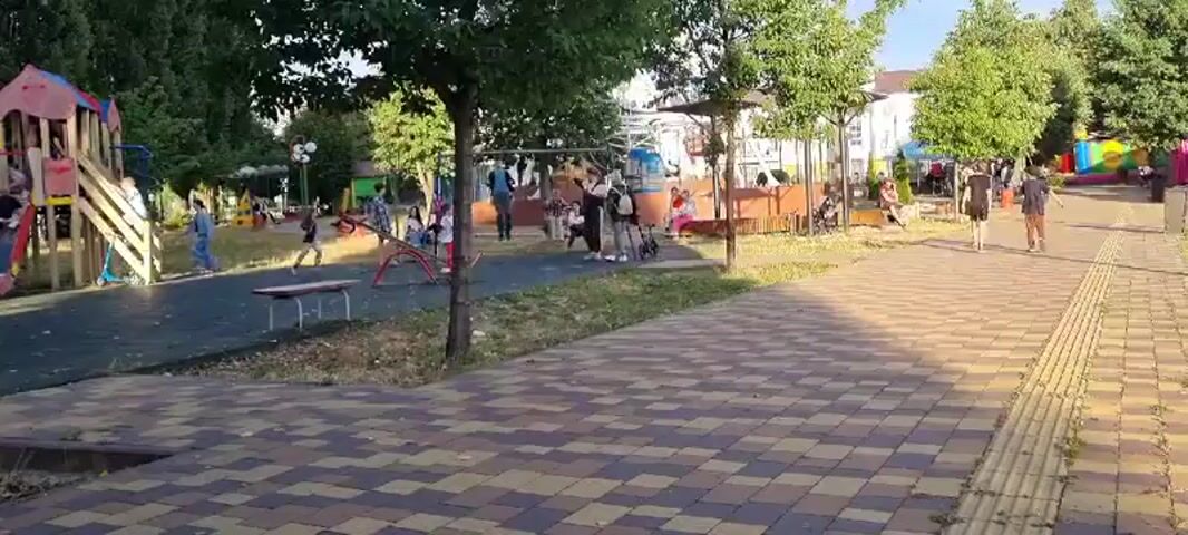 A playground with children playing. There are swings, slides, and other playground equipment. There are trees and benches. The ground is covered with rubber mats and paving stones. The sky is blue and the sun is shining.