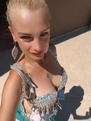 The video shows a blonde woman in a sparkling costume with her hair pulled back. She is looking at the camera and smiling. She is wearing a necklace with a blue pendant and large earrings. She is standing outdoors in front of a building with a tiled roof. In the background, there is a swimming pool with people in it and a bridge. The weather appears to be sunny.