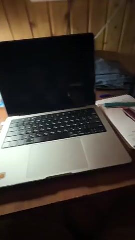 The video pans across a wooden desk. On the desk is a silver laptop with its screen closed, and a keyboard visible. To the left of the laptop is a clear plastic water bottle with a blue label. Further left, partially obscured, is a desk lamp. A white cable is visible near the water bottle and laptop. In the background, a white wall with electrical outlets is visible, and wooden paneling.