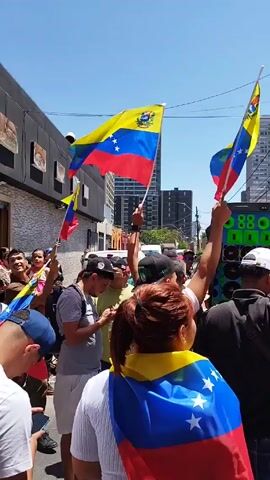 The video shows a daytime street scene with a crowd of people waving Venezuelan flags. Most people are partially visible, with many wearing hats, some are wearing sunglasses and some have flags draped over their shoulders. In the background, there are buildings, a store sign that says "COMERCIALIZADORA MR QUESOS," overhead wires and light poles. A large sound system is visible in the middle of the crowd and music is playing. The people are clapping and appear to be celebrating or protesting. The sky is clear and blue.