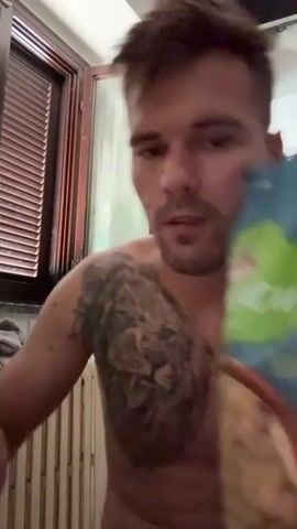 The video shows a man with short brown hair and stubble, shirtless, with a large tattoo of a lion on his chest. He is holding a bag of snacks, which appears to be chips or crackers, with a green and blue design. The man is bringing a snack to his mouth and chewing. He seems to be indoors, in what looks like a kitchen or dining area. There is a window with blinds in the background, and what appears to be a radiator. The lighting suggests it could be daytime. The text on the bag is partially visible and includes the word "SMACK" in large green letters, and some smaller text below it. The overall mood is casual and relaxed.