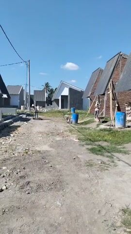 The video shows a construction site with several unfinished houses under a clear blue sky. Two construction workers are visible. One worker, wearing a light-colored shirt and hat, is in the left middle ground, near a pile of dirt and a tool. Another worker, wearing a striped shirt and white helmet, is in the right middle ground, near a wheelbarrow and two blue barrels. The ground is uneven and covered with dirt and gravel. There are construction materials and supplies scattered around the site. The houses have pitched roofs and unfinished exteriors.