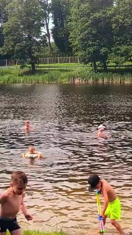 The video shows a group of people, including children, swimming and playing in a lake. In the foreground, a young child is wading in the shallow water near the shore. Behind them, two older children are in the lake, one wearing a black cap and neon green shorts, and the other is partially submerged. In the middle ground, several individuals are swimming in the lake, some with inflatable rings. The background features a lush green treeline and reeds along the edge of the water. The water is rippling, indicating movement.