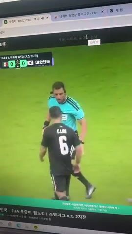 The video shows a computer screen displaying a soccer match being streamed. The scoreboard indicates the match is between Mexico and South Korea, with the score at 0-0. The time is 45:00 and it is the 2nd minute of added time. A referee in a teal jersey is seen interacting with a soccer player in a black jersey with the number 6. Another soccer player in a purple jersey with the number 19 is then shown on screen, appearing to wipe his eye or nose. The background is a green soccer field.