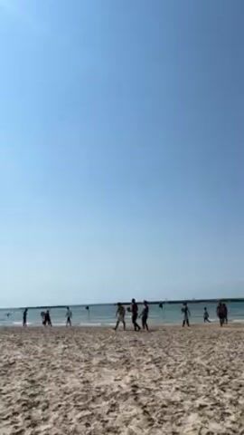 The video captures a beach scene on a sunny day. A group of people, mostly young men, are playing a game on the sand near the water's edge. There is a pier or breakwater visible in the background. The camera focuses on the beach and the players, showing the clear blue sky and calm sea. The sand is textured with footprints.