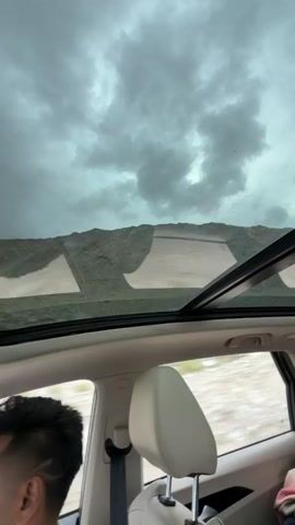 The video begins by showing the view through a car's sunroof. The sky is overcast with dark clouds. The landscape outside consists of barren, rocky mountains. The camera then pans down to show the interior of the car, focusing on the driver, a man with dark hair and a mustache, wearing a white shirt. He is gripping the steering wheel and looking ahead. The car is in motion, and the scenery outside is blurred. The dashboard and infotainment screen are visible. The video then shows the road ahead as the car drives on a winding mountain road. The mountains on the right side of the road are rocky and barren, while on the left, there is a valley with some vegetation. The sky remains cloudy.