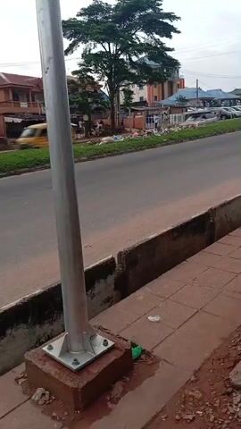 This media file depicts a street scene in Idimu, Nigeria. The foreground is dominated by a metal pole anchored to a concrete base on a tiled sidewalk. Beyond the sidewalk, a paved road runs horizontally across the frame. On the other side of the road, buildings of various heights and colors are visible, including a prominent tree with lush green foliage. Several cars are parked along the roadside, and some appear to be moving, indicated by motion blur. A few people are visible near the buildings. The overall setting suggests a busy, urban environment.

The activity taking place is the movement of vehicles on the road. The time of day appears to be overcast, judging by the diffused light and the appearance of the sky. There are no visible signs or text in the media file. The general atmosphere seems mundane and everyday, with no overt indications of specific events or strong emotions. The presence of some debris along the roadside might suggest a lack of consistent upkeep in certain areas. The concrete barrier between the sidewalk and the road shows signs of wear and tear.