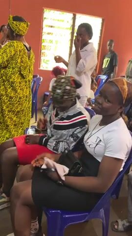 The video shows a group of people sitting and standing in a room with orange walls and windows. Several people are seated on blue plastic chairs, facing forward. One woman in the foreground, wearing a white t-shirt with "perfect" printed on it and a black skirt, is looking towards the right and smiling. To her left, another woman in a white and black patterned jacket and a red skirt is covering her face with her hands. In the background, a woman in a white top and headscarf is standing near a window, appearing to speak or gesture. Other individuals, including men and women of various ages, are visible sitting and standing throughout the room. Some are wearing headscarves and traditional clothing. There is a speaker system visible in the background. The overall atmosphere appears to be a gathering or a meeting.