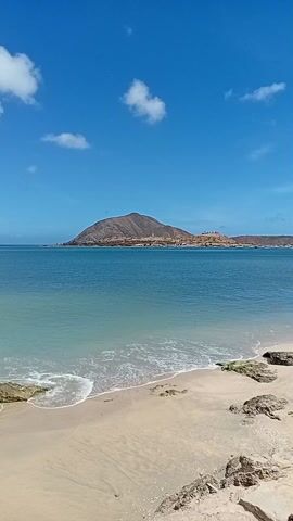 The video shows a beach on a sunny day with a mountain in the background. The water is calm and blue. There are some rocks on the beach and a tree along the edge. The sky is blue with some white clouds. The camera pans to show the length of the beach.