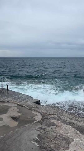The video shows a gray, cloudy day at the seaside. A weathered concrete pier, cracked and worn, stretches out towards the dark, choppy sea. Waves crash against the pier, creating white foam. The ocean stretches to the horizon under an overcast sky. A rocky coastline with some sparse greenery and a single tree is visible in the distance. The overall impression is one of a cold, windy, and slightly desolate coastal scene.