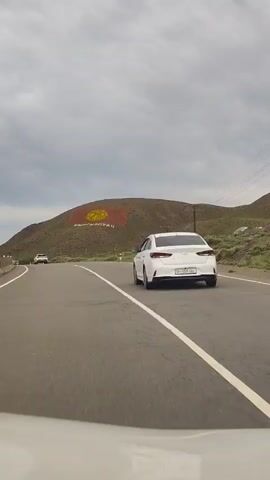 A white car is driving on a paved road. The car is in the foreground and is moving towards the right side of the frame. In the background, there is a large hill with the flag of Kyrgyzstan painted on it. The flag is red with a yellow symbol in the center. The sky is cloudy.