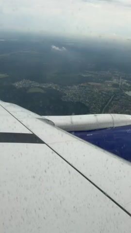 The video captures an aerial view from an airplane window. A white airplane wing, speckled with small dots, dominates the foreground and extends across the lower half of the frame. A blue section of the plane's fuselage is visible on the right side of the wing. Beyond the wing, a landscape unfolds, showing a vast expanse of green forest dotted with numerous houses and buildings, suggesting a suburban or rural area. A body of water, possibly a lake or river, is visible in the distance, along with faint clouds in the sky. The overall scene conveys a sense of travel and perspective from high above the ground.