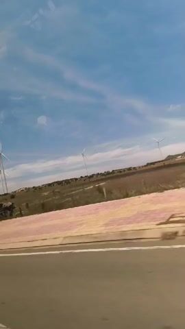 The video is a first-person perspective recording from a vehicle, likely a car or bus, looking out the window. The view is predominantly of a landscape under a blue sky with scattered clouds. In the distance, several wind turbines are visible, suggesting an area with wind energy infrastructure. The immediate foreground features a road or pathway with a textured, light-colored border. To the right of the border, there is an area with several structures, some appearing to be stalls or small buildings with thatched roofs, and various vehicles including a yellow truck and a red truck. The overall impression is of a rural or semi-rural setting, possibly a roadside area with a view of wind farms. There are no people clearly visible in the frames. The light suggests it is daytime, likely sunny.
