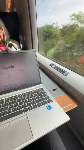 The video shows an interior view of a train carriage, looking out through a large window. In the foreground, a silver HP laptop with an Intel Core i5 sticker is open on a small table next to the window. The laptop screen displays the Google search page. Partially visible to the left is a person's arm and torso, possibly the owner of the laptop, wearing a light-colored sleeve.

Outside the window, a blur of green foliage, including trees and bushes, speeds by, indicating the train is in motion. The sky above the horizon is clear and blue. Reflected in the window is a blurred image of another person, possibly a passenger or a reflection of the person filming, wearing a light green jacket and holding something red. Two black QR code stickers are affixed to the white window frame below the glass. The scene depicts travel on a sunny day through a lush, green landscape, likely in the area of Pyrbaum, Germany, as the train passenger works or browses on their laptop.