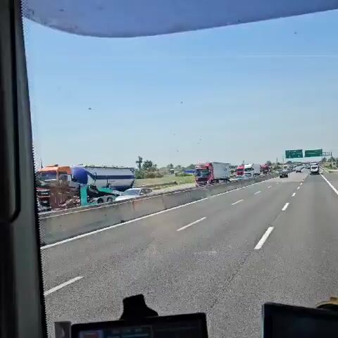The video shows a view from a moving vehicle on a highway. Several large trucks pass by, including a red truck with “DE CERONE” written on the side, carrying what appears to be an excavator. A sign for “Fidenza Village” is visible in the distance. The sky is clear and blue. The road is paved and has a concrete barrier on the side. There is green vegetation in the background.