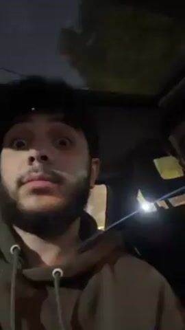 Two men are inside a car at night. The man on the left is looking at the camera with wide eyes and a slightly surprised expression. He has a beard and is wearing a hooded sweatshirt. The man on the right is in the passenger seat, with his eyes closed, seemingly asleep. A sunroof is visible above them, showing trees and the night sky. The car's interior is dark, but there is some light reflecting off the windshield and dashboard. The setting appears to be urban, with streetlights visible in the background.