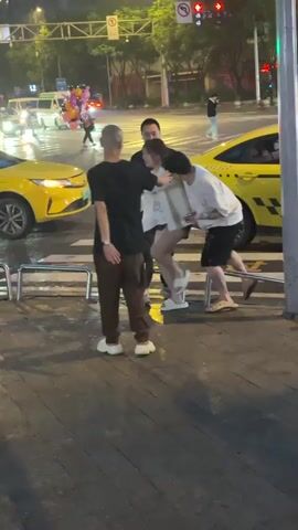 The video captures a street scene at night with several people gathered near a crosswalk. A young woman in a white top and dark shorts is being held by two men, one on her left and one on her right. The man on her left, wearing a black t-shirt and brown pants, has his back to the camera and appears to be pulling her slightly. The man on her right, in a white t-shirt and black shorts, has his arm around her. A fourth man, also in a black t-shirt, is standing next to the man in the white t-shirt, looking towards the woman. Behind them, a yellow taxi is stopped, and another yellow vehicle is partially visible to the left. The street is wet, suggesting it may have recently rained. Traffic lights and streetlights are visible in the background, illuminating the scene. There are also balloons in the background, held by an unseen person. The overall impression is of a tense or potentially confrontational interaction.