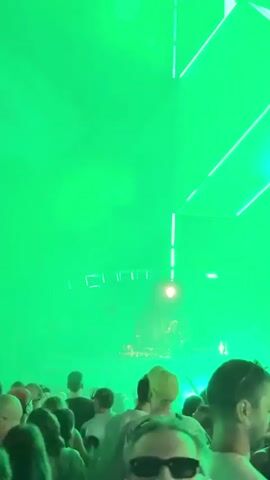 A large crowd is gathered at an outdoor nighttime event, likely a music festival or concert. The stage is illuminated by bright lights that change color, cycling through red and green. A DJ, identifiable by a female figure with red hair, is behind a DJ booth at the center of the stage, appearing to be mixing music. The crowd is densely packed, with many individuals holding up their phones to record the performance. The overall atmosphere is energetic, with the lighting and the crowd's engagement suggesting a lively party or festival environment.