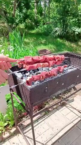 This is a close-up, medium shot video of several skewers of raw meat being grilled over hot coals in a rusty barbecue grill. The scene is set outdoors in a garden or backyard area, with lush green foliage and trees visible in the background. The sun appears to be shining, indicating it is daytime, likely late morning or early afternoon, with pleasant weather. The grill is made of metal and has a rustic, well-used appearance. The meat is cut into cubes and threaded onto metal skewers, with some skewers already placed on the grill and others being added by an unseen person whose hands are partially visible on the left side of the frame. The coals are glowing red and white, suggesting they are very hot and ready for grilling. The overall impression is one of a casual outdoor cooking event, perhaps a barbecue or picnic. No visible text is present in the video. The location context is identified as Vyazovka, Russia.