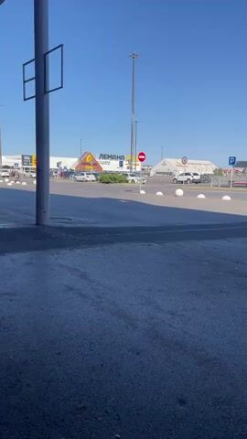 The video shows a person riding a dirt bike in a parking lot or open area. The person is wearing a helmet and riding gear. They are riding in a straight line across the paved area. In the background, there are buildings, cars, and a large pole with lights. The sky is clear and blue. There are also white spherical bollards in the foreground and mid-ground.