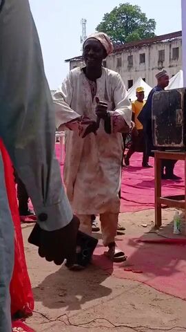 A video captures a man in traditional clothing, a flowing white robe and pants, standing outdoors on a dirt surface with a pink carpet laid out. He is wearing sandals and a headwrap. In his left hand, he holds a cane, and in his right hand, he gestures animatedly towards his face, possibly speaking into something not clearly visible or fanning himself. Behind him, other people are visible, some appearing to be security personnel in dark uniforms, and others in the background near tents and buildings. The scene appears to be an outdoor event or gathering, with the man as the central focus of the frame.