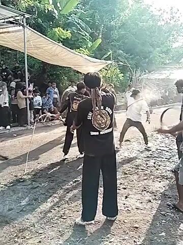 The video shows a group of people engaged in what appears to be a traditional combat sport or performance. Several people are seen wielding what look like flexible weapons, possibly made of rope or vine with weights attached to the end. They are swinging these weapons at each other in a display of skill and agility.  The environment is an outdoor space, possibly a village road or square, with a canopy or tent-like structure visible in the background. There is a crowd of people gathered, watching the event. Some of them appear to be seated under the canopy. The ground appears to be muddy or wet. The atmosphere seems lively and festive, with a sense of cultural celebration.