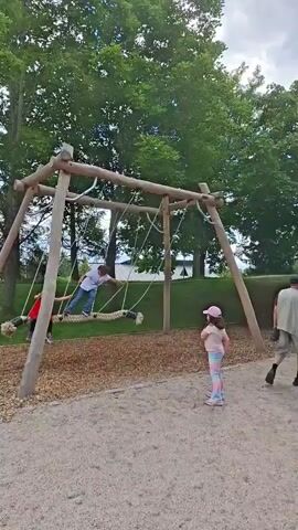 A group of children are playing on a wooden swing set in a park. Two children, one wearing a red dress and the other a blue shirt, are on the swings, while another child in a pink hat and striped leggings watches from the ground. The swing set is made of natural wood and is covered with wood chips. Trees and green grass are visible in the background.