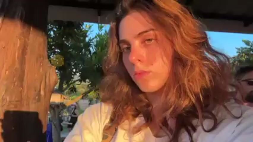 A woman with wavy brown hair is standing outdoors in a warm, golden light. She is wearing a white t-shirt and has what appears to be a strap or suspender over her shoulder. The camera angle is a selfie, looking slightly down. The video moves, showing other people around her, standing on what appears to be a stone or concrete surface. Their feet are visible; some are wearing sandals. The environment seems to be an outdoor gathering place, potentially a patio or yard, with trees and possibly buildings visible in the background.