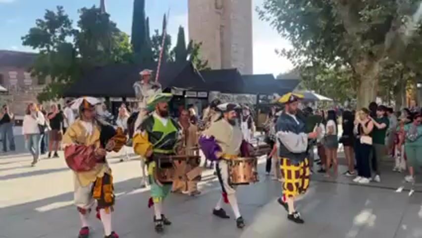A group of costumed performers parade in a public square. Four men wearing colorful Renaissance-style clothing march while playing hand-held drums. Behind them, a person in full medieval armor rides a white horse. The group parades through a crowd of onlookers on a sunny day. Trees and a tall brick structure can be seen in the background, along with vendor stalls.