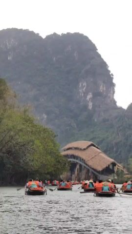 The video shows a number of boats on a river, all filled with people wearing orange life vests. They are rowing towards a building with a thatched roof that spans the river. In the background, there are large, forested mountains under a cloudy sky.