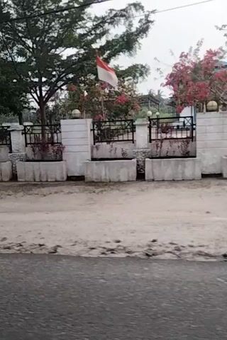 The video pans from a building with a balcony and some potted plants to an outdoor scene. The scene includes a grassy area with some trees and a sign that appears to be for sale or rent. There is also a road or path in the foreground, and the overall impression is of an undeveloped or partially developed area. The weather appears to be overcast.