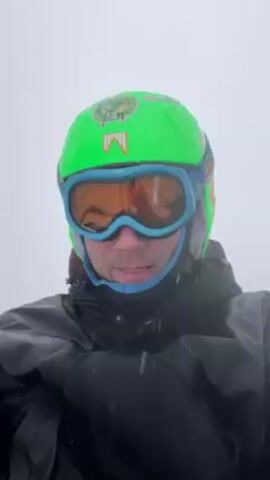 A person wearing ski gear sticks their tongue out at the camera. The person is wearing a green ski helmet with a Marlboro logo sticker, blue-rimmed ski goggles with orange lenses, and a black jacket. Snow is falling, and the background is an overcast sky.
