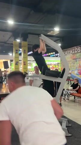 A young male with dark hair and glasses, wearing a black t-shirt and black pants, is performing pull-ups on a white and gray pull-up bar machine. He is suspended from the bar with his body hanging down, then pulling himself up. He repeats this motion. A man in a white t-shirt and blue jeans stands below him, looking up. Another person in a white t-shirt is partially visible in the foreground, walking away from the camera. The scene appears to be indoors in a public space, possibly a mall or fitness expo, with bright lighting and banners in the background. Some people are seated on a bench to the right, observing the activity.
