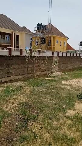 The video shows a wide shot of a grassy, sandy area with buildings in the background. A low brick wall runs along the left side of the frame, separating the grassy area from the properties behind it. There are two houses visible; one painted light yellow with a brown roof and another painted a paler yellow with a darker brown roof. Both houses have water tanks on metal stands. A tall radio tower is visible behind the yellow house. The sandy area has patches of green grass and small plants growing throughout. In the distance, more houses and trees can be seen under a light blue sky. The camera pans slowly to the right, revealing more of the sandy area and distant buildings.  The terrain is mostly flat.