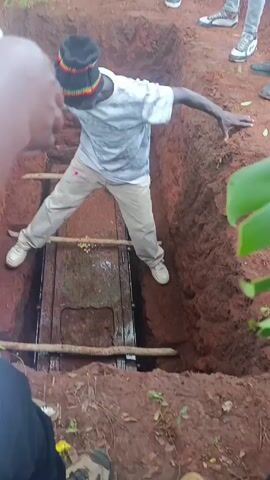 The video shows a rectangular, metal coffin resting at the bottom of a dug grave. Three wooden sticks are placed across the coffin horizontally. A man wearing a white t-shirt, light-colored pants, white shoes, and a black beanie with red, yellow, and green stripes stands inside the grave, on top of the coffin, balancing with his arms outstretched to the sides for stability. The grave is surrounded by reddish-brown soil.  Several pairs of shoes and lower legs of people are visible around the perimeter of the grave. A few green leaves are visible at the right edge of the frame.