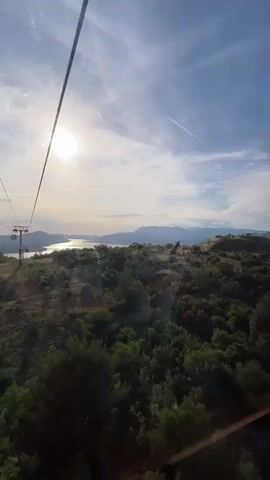 A person is riding in a cable car looking out at the scenery. The cable car is moving and the scenery outside changes from rolling hills and forests to a view of a body of water with a town on its banks. Mountains are visible in the distance. The person is wearing a white top, blue jeans, and a striped shirt. They have sunglasses on their head and are looking out the window. A backpack is on the seat next to them.