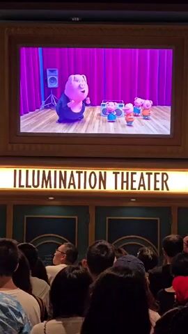 This media file captures a scene in front of the "ILLUMINATION THEATER" in Tongzhou District, China. The focus is on a large screen displaying an animated scene featuring several pig characters. A larger pig in a sparkly black outfit is on the left, with its arms raised. To the right, three smaller pigs in blue and orange outfits are dancing. A boombox sits on the wooden stage. The background features purple curtains.

Below the screen, the illuminated sign "ILLUMINATION THEATER" is prominent. In front of the theater entrance, a crowd of people are gathered, facing the screen and seemingly watching the animation. The people are diverse in age and gender, and their backs are mostly to the camera. The overall atmosphere appears to be one of entertainment and enjoyment, possibly at an amusement park or a movie theater. The lighting suggests it is likely evening or indoors.