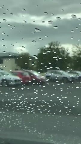 The video shows a close-up view of raindrops on a window, with a blurry background of a parking lot. Several cars are visible in the parking lot, including a red car and several silver or white cars. The sky appears overcast and gray. The focus of the video is the raindrops distorting the view of the scene behind the window. The camera is stationary. The lighting is dim, consistent with an overcast day. The overall atmosphere is subdued and perhaps melancholic due to the rainy weather.